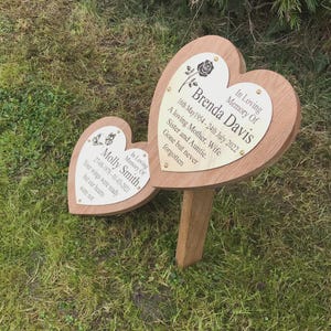 May include: Two heart-shaped wooden memorial plaques with engraved text. One reads "Brenda Davis" with dates and a rose graphic. The other reads "Molly Smith" with dates and butterfly graphics. Both plaques are mounted on a wooden stake for outdoor use.