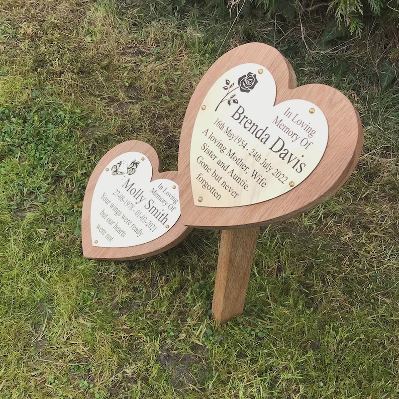 May include: Two heart-shaped wooden memorial plaques with engraved text. One reads "Brenda Davis" with dates and a rose graphic. The other reads "Molly Smith" with dates and butterfly graphics. Both plaques are mounted on a wooden stake for outdoor use.