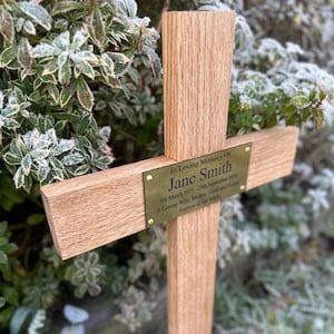 May include: A wooden memorial cross with a brass plaque. The plaque is inscribed with "In Loving Memory Of Jane Smith" and dates. The cross is set against a backdrop of green and frosted foliage.