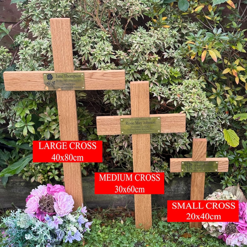 May include: Three wooden memorial crosses with brass nameplates, set in a garden. The largest cross is 40x80cm, the medium cross is 30x60cm, and the smallest cross is 20x40cm. The crosses are surrounded by greenery and flowers.