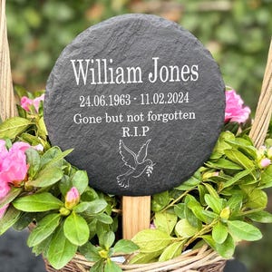May include: A circular, dark grey slate memorial plaque inscribed with the name "William Jones" in white. The plaque features dates, the phrase "Gone but not forgotten," and a dove illustration. It is set in a basket of pink flowers and green foliage.
