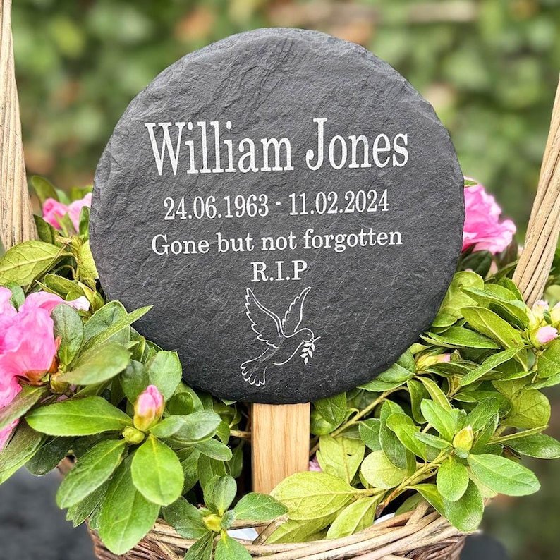 May include: A circular, dark grey slate memorial plaque inscribed with the name "William Jones" in white. The plaque features dates, the phrase "Gone but not forgotten," and a dove illustration. It is set in a basket of pink flowers and green foliage.
