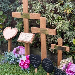 May include: A collection of wooden memorial crosses and slate plaques in a garden. Each cross has a brass nameplate. Heart and oval-shaped slate plaques with inscriptions and floral arrangements are placed in front of the crosses. The text includes names and dates.