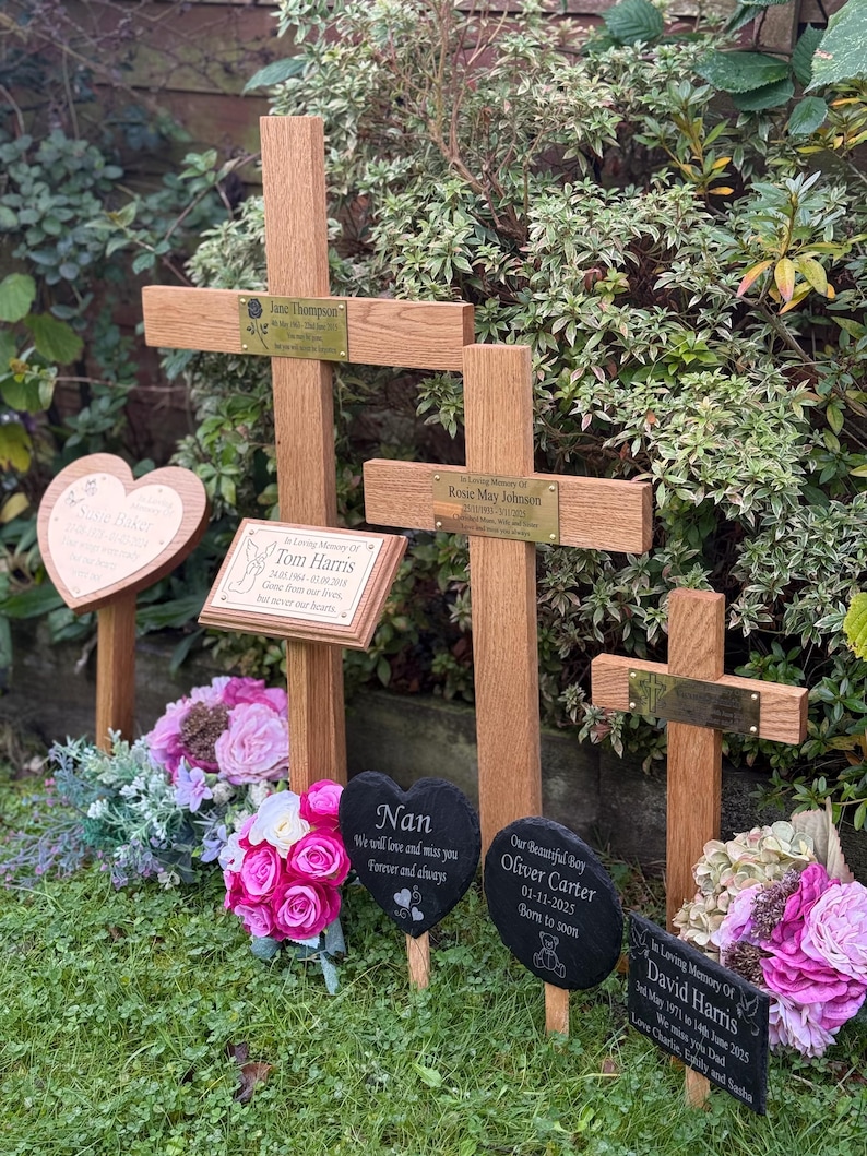May include: A collection of wooden memorial crosses and slate plaques in a garden. Each cross has a brass nameplate. Heart and oval-shaped slate plaques with inscriptions and floral arrangements are placed in front of the crosses. The text includes names and dates.