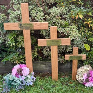 May include: Three wooden memorial crosses of different sizes, each with a name plaque, stand in a garden. The crosses are surrounded by green grass and floral arrangements in shades of pink and purple, creating a peaceful memorial.