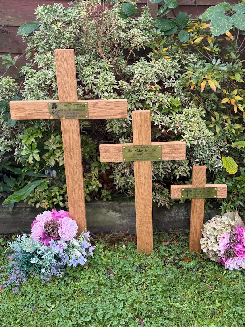 May include: Three wooden memorial crosses of different sizes, each with a name plaque, stand in a garden. The crosses are surrounded by green grass and floral arrangements in shades of pink and purple, creating a peaceful memorial.