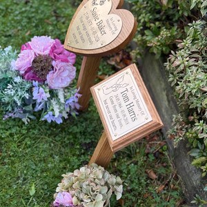 May include: Two wooden memorial plaques with brass plates. One is heart-shaped, inscribed "Susie Baker" with dates and a quote. The other is rectangular, inscribed "Tom Harris" with dates and a quote. They are set in grass with floral arrangements.