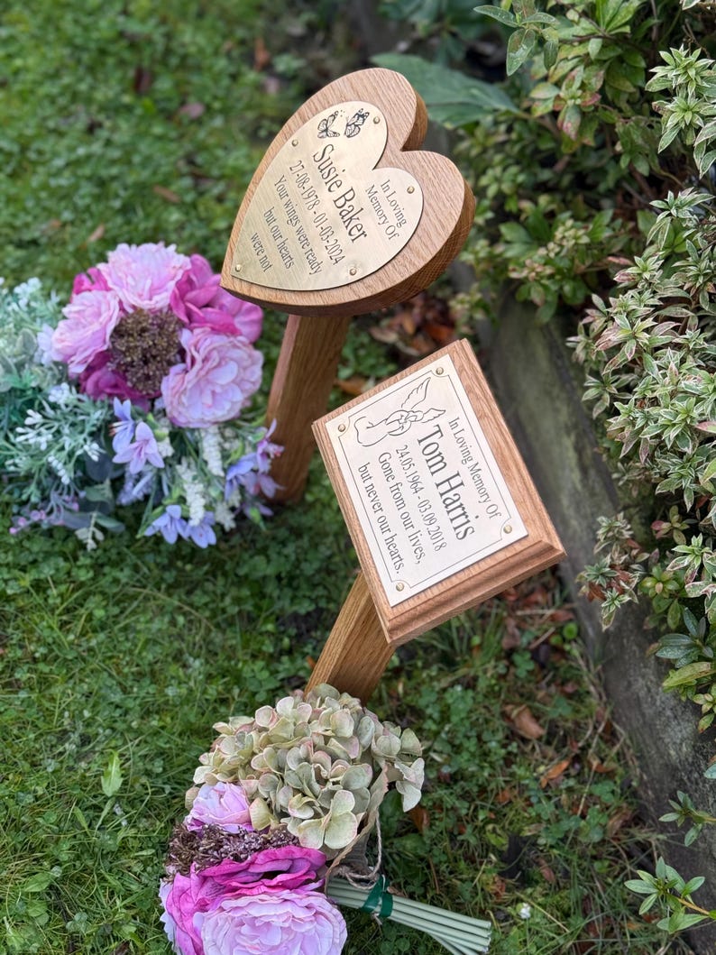 May include: Two wooden memorial plaques with brass plates. One is heart-shaped, inscribed "Susie Baker" with dates and a quote. The other is rectangular, inscribed "Tom Harris" with dates and a quote. They are set in grass with floral arrangements.