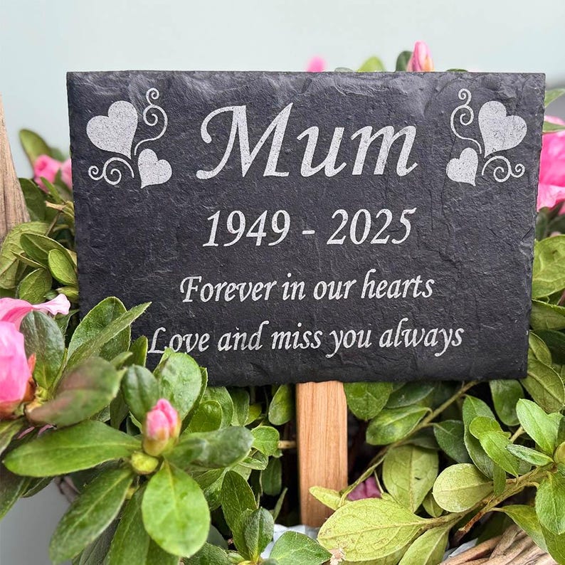 May include: A rectangular black slate memorial plaque with the word "Mum", dates 1949-2025, and the inscription "Forever in our hearts, Love and miss you always" in white. The plaque features heart designs and is surrounded by green leaves and pink flowers.