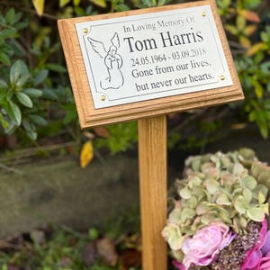 May include: A wooden memorial plaque with the text "In Loving Memory Of Tom Harris 24.05.1964 - 03.09.2018 Gone from our lives, but never our hearts." The plaque features an angel illustration and is mounted on a wooden stake.