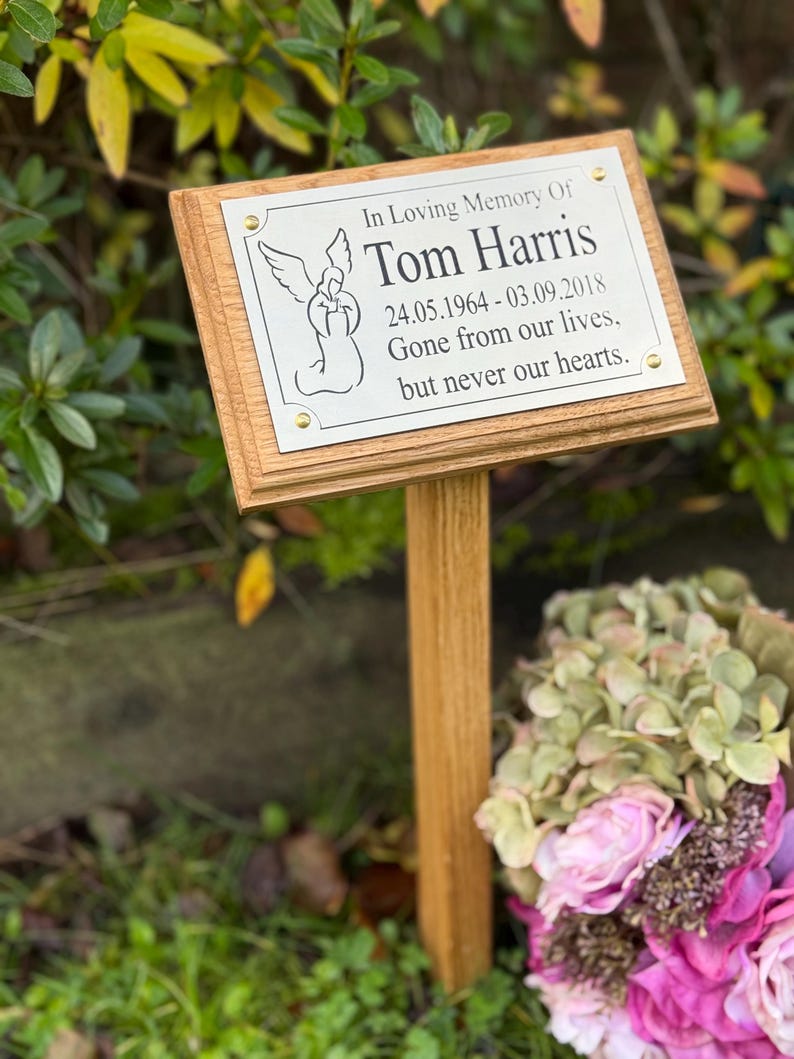 May include: A wooden memorial plaque with the text "In Loving Memory Of Tom Harris 24.05.1964 - 03.09.2018 Gone from our lives, but never our hearts." The plaque features an angel illustration and is mounted on a wooden stake.
