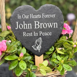 May include: A heart-shaped slate memorial plaque with the inscription "In Our Hearts Forever John Brown Rest In Peace." The plaque is set in a woven basket filled with pink flowers and green foliage.