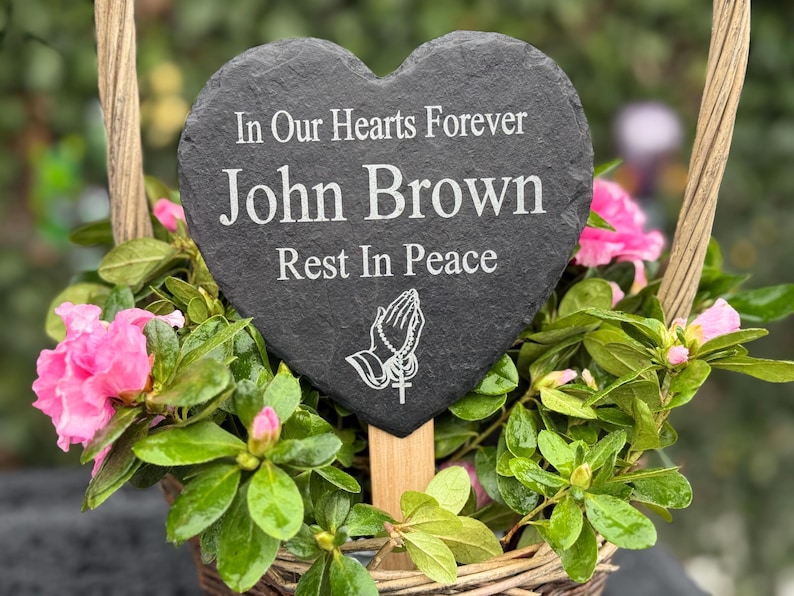 May include: A heart-shaped slate memorial plaque with the inscription "In Our Hearts Forever John Brown Rest In Peace." The plaque is set in a woven basket filled with pink flowers and green foliage.