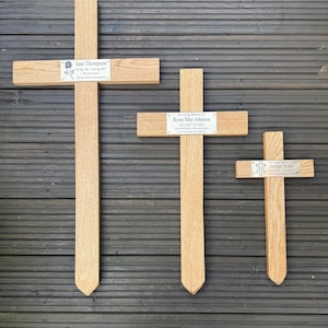 May include: Three wooden memorial crosses of different sizes. Each cross features a metal plaque with a name and dates. The largest cross reads "Jane Thompson". The middle cross reads "Rosie May Johnson". The smallest cross reads "George Smith".