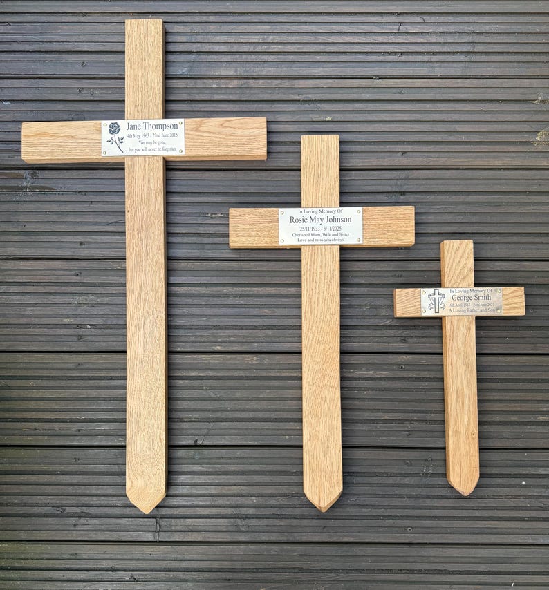 May include: Three wooden memorial crosses of different sizes. Each cross features a metal plaque with a name and dates. The largest cross reads "Jane Thompson". The middle cross reads "Rosie May Johnson". The smallest cross reads "George Smith".