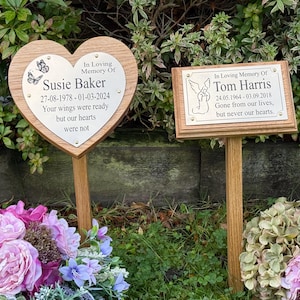 May include: Two memorial plaques on wooden stakes. One is heart-shaped with the text "In Loving Memory Of Susie Baker" and dates. The other is rectangular with "In Loving Memory Of Tom Harris" and dates. Both have engraved text.