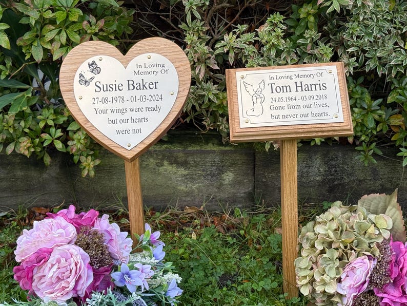 May include: Two memorial plaques on wooden stakes. One is heart-shaped with the text "In Loving Memory Of Susie Baker" and dates. The other is rectangular with "In Loving Memory Of Tom Harris" and dates. Both have engraved text.
