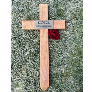 May include: A wooden cross with a plaque inscribed "In Loving Memory Of Jane Smith." A red rose is placed on the cross. The plaque includes dates and a dedication. The cross is set on frosted grass.