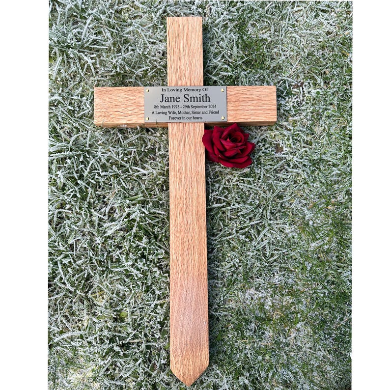 May include: A wooden cross with a plaque inscribed "In Loving Memory Of Jane Smith." A red rose is placed on the cross. The plaque includes dates and a dedication. The cross is set on frosted grass.