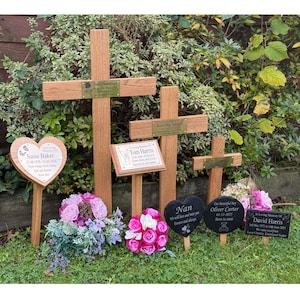 May include: A selection of wooden memorial crosses and plaques, some with floral arrangements. The crosses have brass nameplates with names and dates. Heart-shaped and oval plaques are also present, with text such as "Susie Baker" and "Nan."
