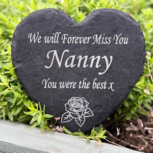 May include: A heart-shaped, dark grey slate memorial stone. The inscription reads "We will Forever Miss You Nanny You were the best x" in white. A rose illustration is at the bottom. The stone is surrounded by green foliage.