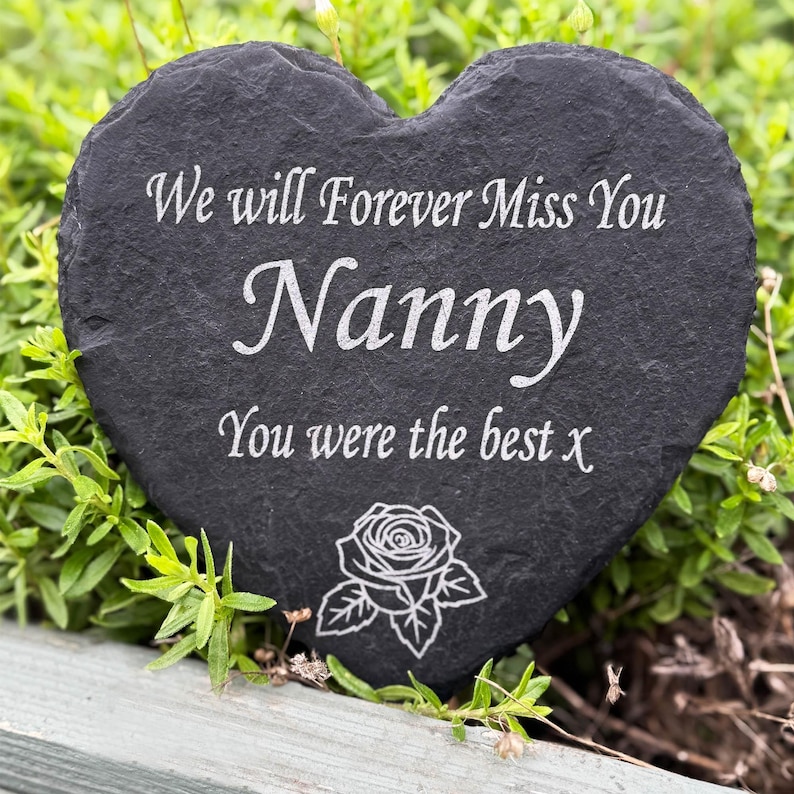 May include: A heart-shaped, dark grey slate memorial stone. The inscription reads "We will Forever Miss You Nanny You were the best x" in white. A rose illustration is at the bottom. The stone is surrounded by green foliage.