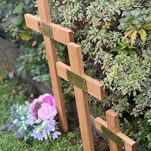 May include: Three wooden crosses of different sizes, each featuring a brass plaque. They stand in a grassy area, with green and variegated foliage in the background. A small bouquet of pink and purple flowers is placed nearby.