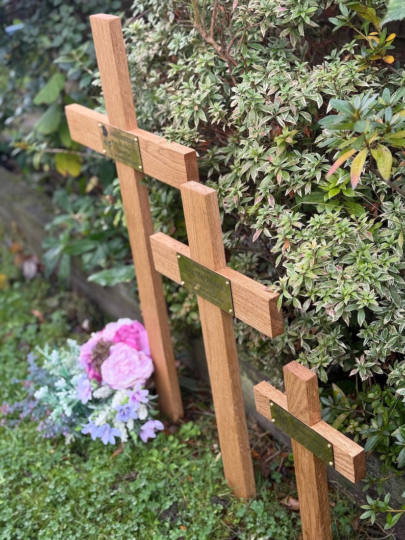 May include: Three wooden crosses of different sizes, each featuring a brass plaque. They stand in a grassy area, with green and variegated foliage in the background. A small bouquet of pink and purple flowers is placed nearby.