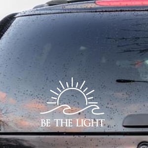 May include: White vinyl decal featuring a sun rising over ocean waves, with the text "BE THE LIGHT" below. The decal is applied to the rear window of a vehicle, suggesting a positive and inspirational theme.