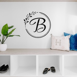 May include: A black and white wall decal featuring the letter "B" inside a circular design with a leafy accent. The decal is mounted on a white wall above a built-in shelf with decorative pillows, a ceramic jug, and shoes.