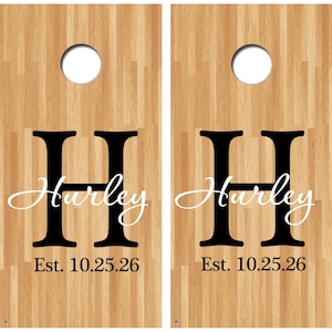 May include: Two wooden cornhole boards with a light brown wood grain pattern. Each board features a large black letter "H", the name "Hurley" in a cursive font, and the date "Est. 10.25.26" in black. A white circular hole is at the top.