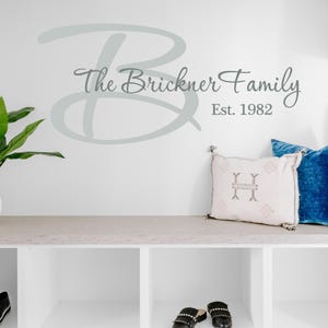 May include: A wall decal with the text "The Brickner Family" and "Est. 1982" in a script font. A large letter "B" is above the text. Two decorative pillows and a plant are on a shelf below the decal. Black shoes are on the shelf.