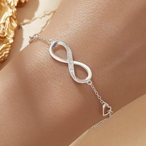 Peut inclure: Un bracelet argenté orné d'un symbole infini serti de petites pierres scintillantes. Le bracelet est relié par une fine chaîne, avec un petit cœur à l'extrémité. Le logo Bella Finn est visible en haut à droite.