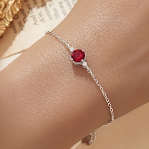 Könnte beinhalten: Ein filigranes Silberarmband mit einem leuchtend roten Edelstein als Mittelpunkt. Das Armband hat eine feine Kette und ist mit kleinen, funkelnden Akzenten auf beiden Seiten des Hauptsteins verziert. Das Bella Finn-Logo ist sichtbar.