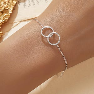 May include: A delicate silver bracelet featuring two interlocking circles. The circles have a textured design and are connected by a fine chain. The bracelet is displayed on a wrist, showcasing its elegant simplicity.