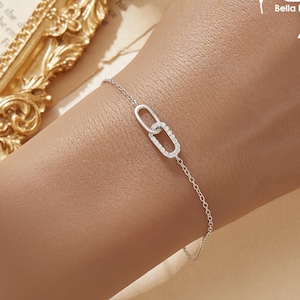 May include: A delicate silver bracelet featuring two interlocking oval links, one of which is encrusted with small, sparkling crystals. The bracelet is shown on a wrist, with a subtle, elegant design.