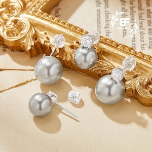 以下が含まれることがあります： シルバーグレーのパールイヤリング。クリアな正方形のクリスタルアクセント付き。イヤリングは、背景に金色の装飾フレームがある、明るいベージュの表面に表示されています。クラシックなジュエリーアイテムです。