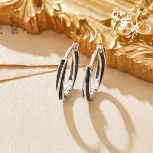 Op de afbeelding: Een paar zilveren oorringen met een zwarte emaille streep. De oorbellen worden gepresenteerd op een crèmekleurig oppervlak met een goudkleurig decoratief frame op de achtergrond. De oorbellen hebben een eenvoudig, elegant ontwerp.