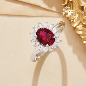 May include: A silver ring featuring a large, oval-cut ruby gemstone surrounded by a halo of smaller, round diamonds. The ring's band is slender and polished, with the Bella Finn logo visible.