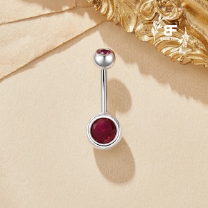 May include: A silver belly button ring with a polished finish. It has a small, round, deep red gemstone at the top and a larger, matching gemstone at the bottom. The gemstones are set in a silver bezel.