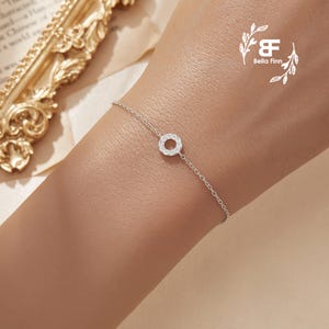 May include: A delicate silver bracelet with a small circle charm encrusted with tiny clear stones. The bracelet is on a person's wrist.