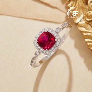 Può includere: Un anello d'argento con una gemma rossa intensa di forma quadrata, circondata da un alone di pietre chiare più piccole. La fascia presenta un design delicato con ulteriori piccole pietre. L'anello è posto su uno sfondo neutro.