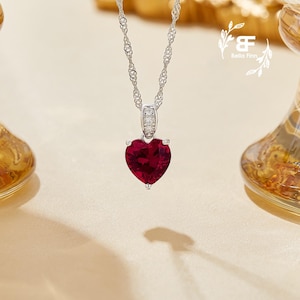 May include: A silver necklace with a heart-shaped pendant featuring a large, deep red gemstone. The pendant is accented with smaller, clear gemstones.