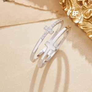 以下が含まれることがあります： 十字架のデザインが施された2つのシルバーリング。1つのリングには、小さな輝く石で飾られた十字架があります。もう一方のリングには、無地の白い十字架があります。リングはニュートラルな背景に表示されています。