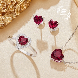 Peut inclure: Un ensemble de bijoux en forme de cœur comprenant une bague, des boucles d'oreilles et un collier. Chaque pièce présente une pierre précieuse rouge entourée d'un halo de petites pierres blanches. Les bijoux sont sertis en argent.