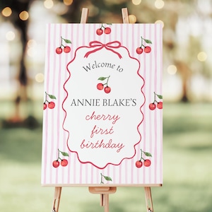 Puede incluir: Un letrero de bienvenida para una fiesta de primer cumpleaños. El letrero presenta un fondo a rayas rosas y blancas con ilustraciones de cerezas. El texto "Welcome to ANNIE BLAKE'S cherry first birthday" está escrito en una fuente decorativa, enmarcado por un borde rojo.