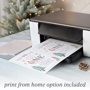 May include: A white and black home printer outputting two identical rectangular paper invitations. The invitations feature pink and green holiday-themed graphics, including a Christmas tree, ornaments, and the text Our Little Sugarplum Is Turning ONE.