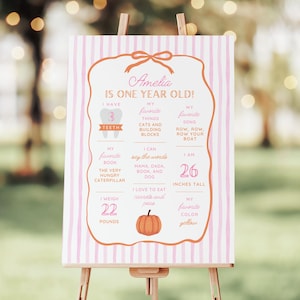 Puede incluir: Un letrero a rayas rosas y blancas sobre un caballete de madera. El letrero tiene el texto "¡Amelia tiene un año!" e incluye detalles sobre la edad, la altura (66 cm), el peso (10 kg) y las cosas favoritas del niño.