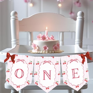 Könnte beinhalten: Ein weißer Hochstuhl trägt einen kleinen Kuchen, der mit rosa Blüten und einer brennenden Kerze verziert ist. Darunter hängt ein Banner mit dem Wort "ONE" in roten Buchstaben. Das Banner hat einen rosa-weiß gestreiften Hintergrund mit Kirschillustrationen und roten Schleifen.