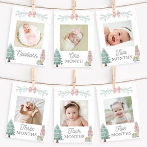 May include: Six rectangular milestone cards hanging on twine with wooden clips. Each card features a baby photo, a pink bow, a Christmas tree, a nutcracker, and text indicating age from Newborn to Five Months in a script and block font style.